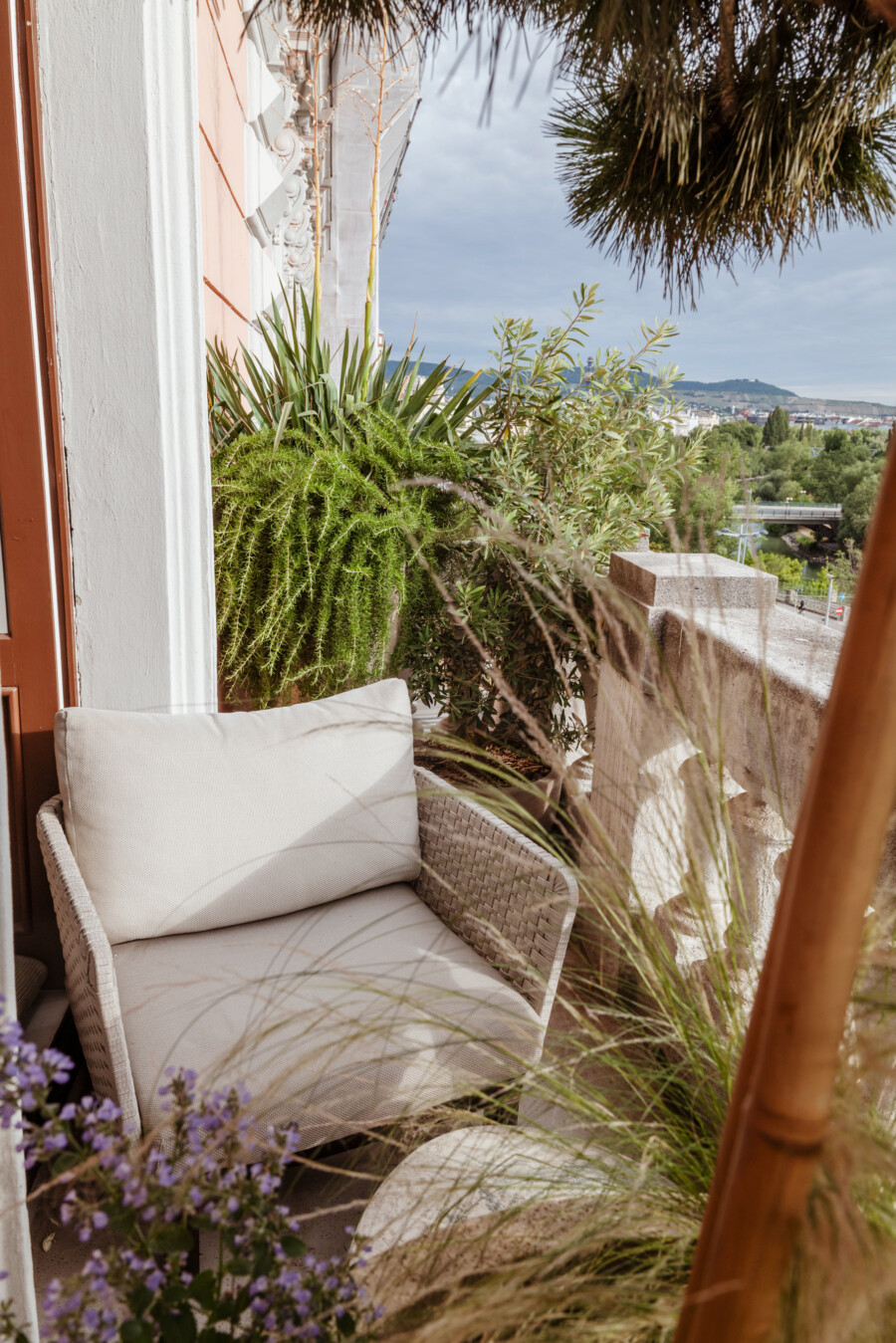 Balkonstuhl aus Geflecht mit hellen Kissen; Lavandula angustifolia (Lavendel) und Pennisetum setaceum; Yucca sp.; Stadtblick im Hintergrund.