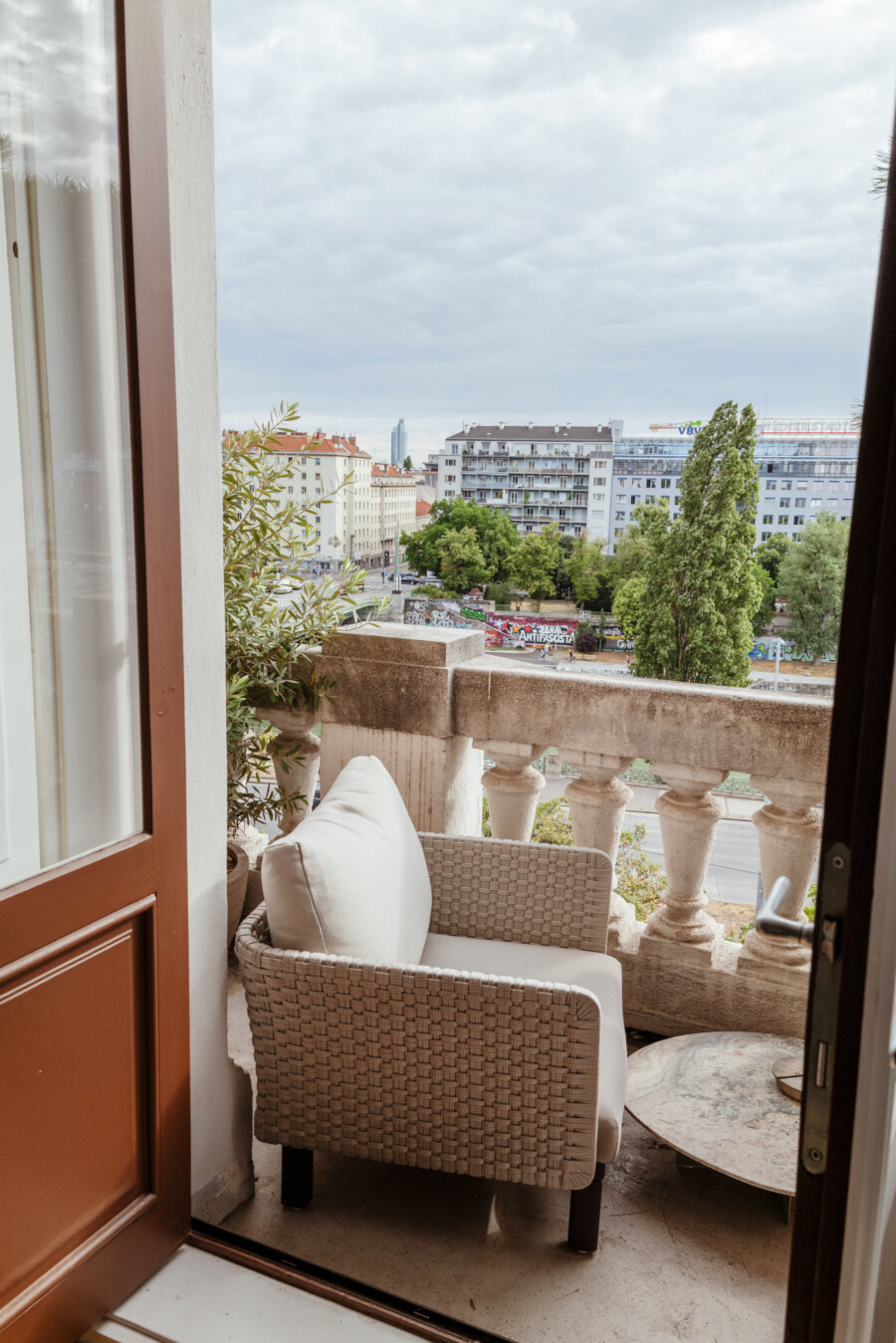 Geflecht-Sessel auf Balkon; Olivenbaum (Olea europaea) im Vordergrund, städtische Häuserlandschaft mit Bäumen im Hintergrund.