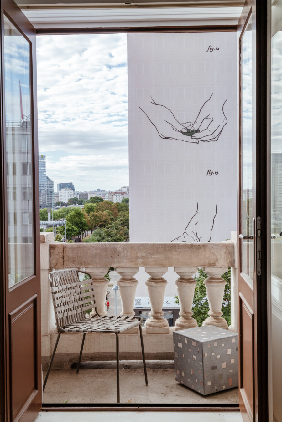 Balkon mit Metallstuhl vor Balustrade; Stadtlandschaft mit Bäumen im Hintergrund; Wandillustration zeigt Hände, die etwas halten.