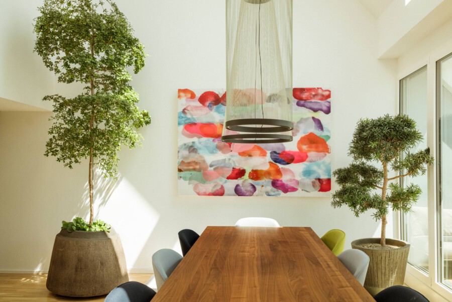 Heller Konferenzraum mit Holzkonferenztisch und Pendelleuchte; links Ficus benjamina, rechts Chamaecyparis pisifolia; abstrakte Wandmalerei.