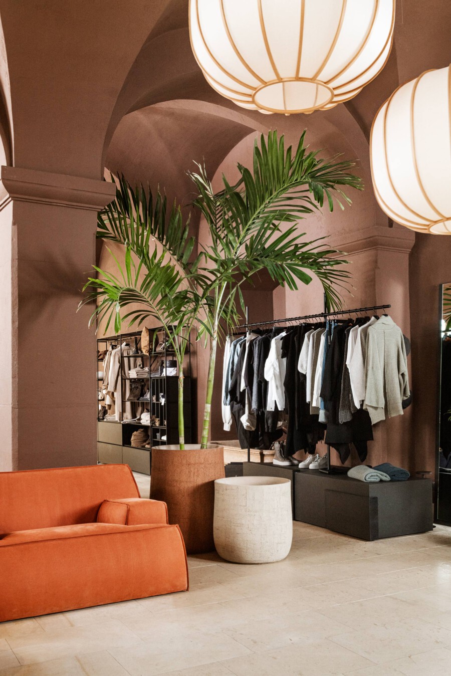 Innenansicht einer Modeboutique mit Kleiderstange; mehrere Dypsis lutescens-Palmen in Terrakotta-Töpfen; orange Sitzgelegenheiten und runde Lampen.
