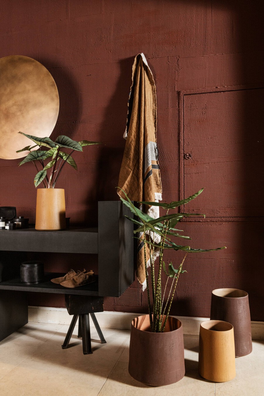 Wohnzimmerdeko vor roter Ziegelwand; Zamioculcas zamiifolia (ZZ-Pflanze) im orangefarbenen Topf, dunkler Tisch mit Schuhen, Tuch hängt an Wand.