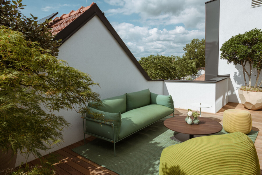 Dachterrasse mit Holzdeck, grünem Sofa, rundem Couchtisch, gelb-grüner Pouf, Topfpflanzen und weißen Wänden; blauer Himmel mit Wolken.