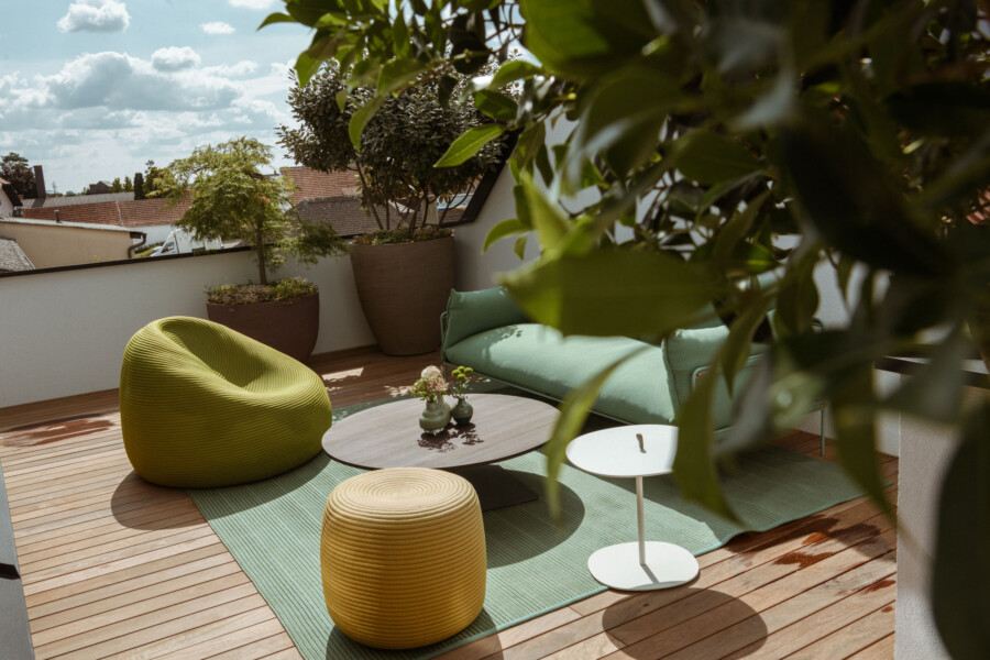 Holz-Terrasse mit grüner Lounge, gelben Poufs, rundem Tisch und weißem Beistelltisch; große Pflanzkübel, grüne Matte, von Blättern umrahmt.