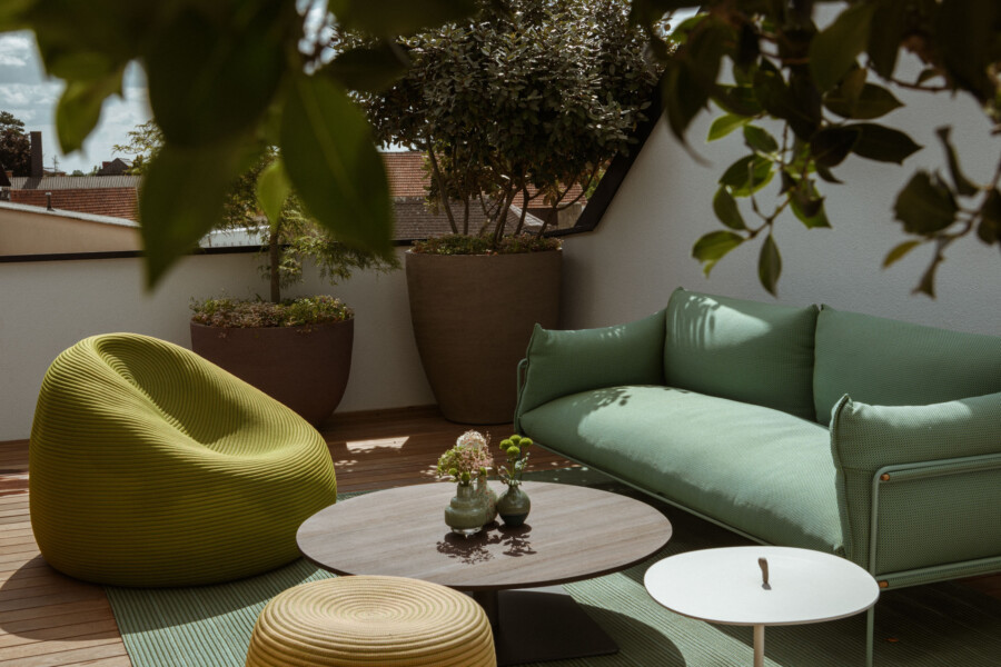 Auf einer sonnigen Dachterrasse eine mintgrüne Sofa-Lounge, ein gelber runder Sitzsack, ein runder Holztisch, kleine Vasen und große Pflanzkübel.