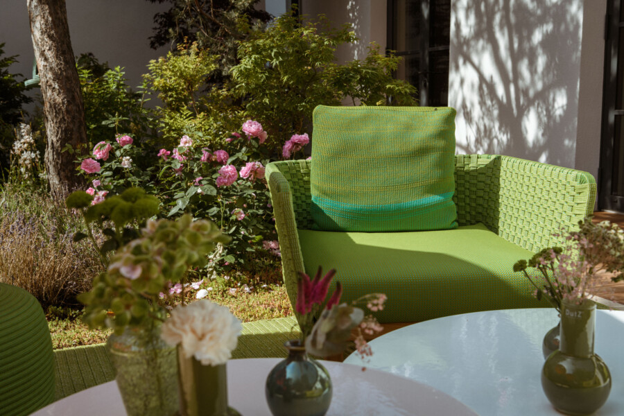 Grüner Korbwaren-Sessel im Garten mit Kissen; umliegende rosa Rosen, Grünpflanzen und Schatten an der weißen Wand; runder weißer Tisch mit Vasen im Vordergrund.