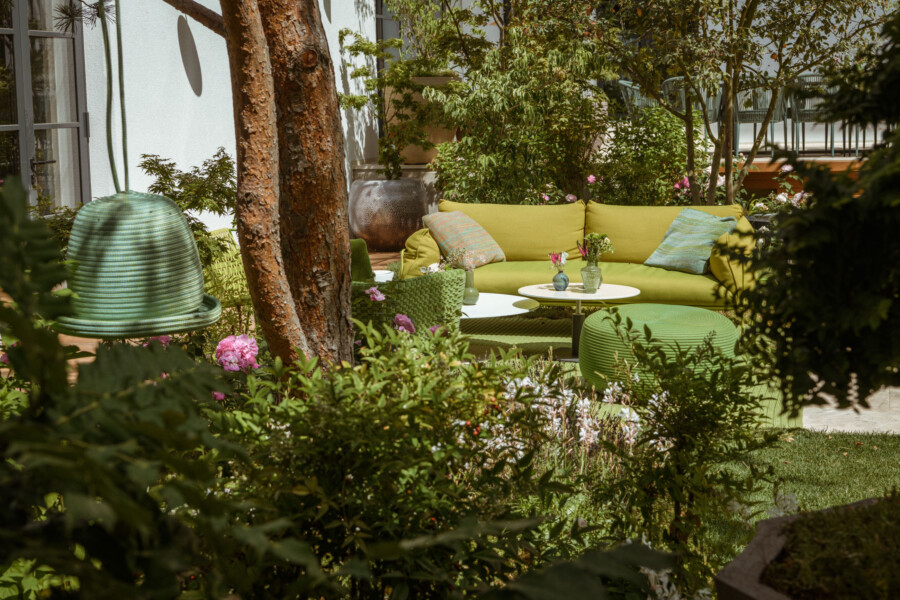 Gartenlounge mit gelber Couch, grünen Kissen, runden Beistelltischen und viel Grün; links ein großes grün geripptes Outdoor-Objekt neben einem Baumstamm.