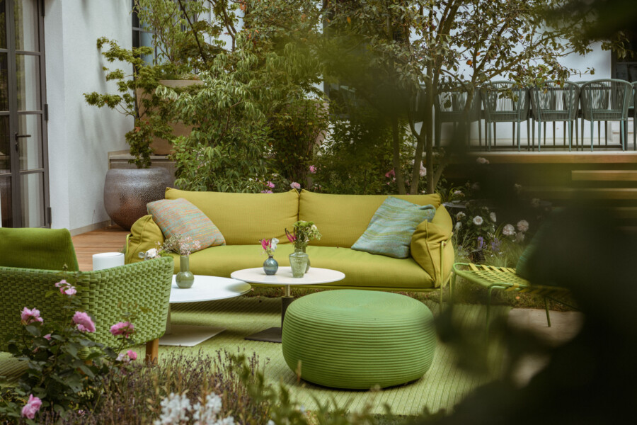 Gartenlounge mit gelbgrüner Couch, bunten Kissen und runden weißen Tischen; grünem Pouf, umgeben von Pflanzen, Balkon mit Stühlen dahinter.