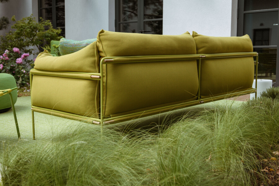 Gelbgrünes Garten-Sofa mit Metallrahmen auf Grasboden vor einer Hausfassade; links rosa Rosen und viel Grün.