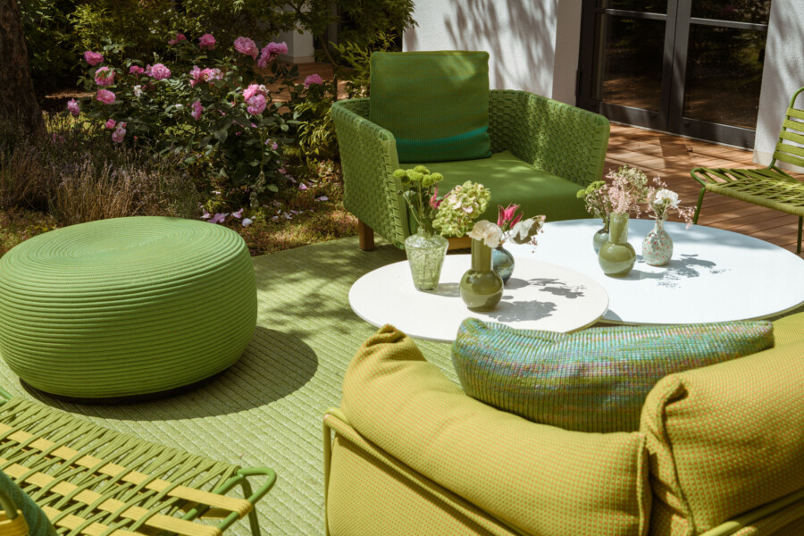 Grüne Gartenmöbel auf Terrasse: runder weißer Tisch mit Blumenvasen, gepolsterte Korbsessel, großer grüner Pouf, weitere grüne Stühle, Rosen im Hintergrund.