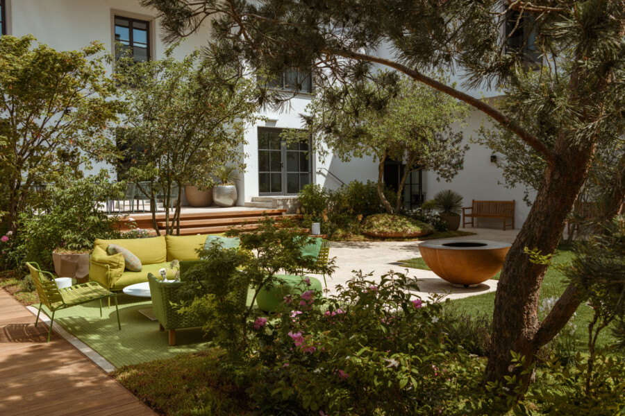 Gartenhof vor einem weißen Gebäude, grüne Lounge-Möbel auf grünem Teppich, bunte Blumen, großer Baum, orange runder Behälter mit Innenraum, Holzbank.