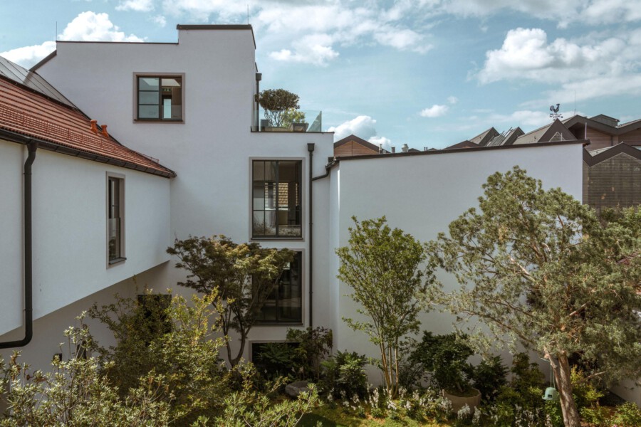 Moderner weißer Gebäudekomplex mit Innenhof, großen Fenstern und Glasfronten; umlenkender Garten mit Bäumen und Sträuchern, blauer Himmel und Wolken.