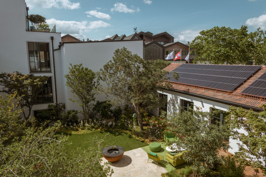 Heller Innenhof mit weißen Gebäuden, Solarzellen auf dem Dach, grünem Garten, zentralem Baum und Lounge-Ecke mit grünen Kissen; Flaggen auf dem Dach