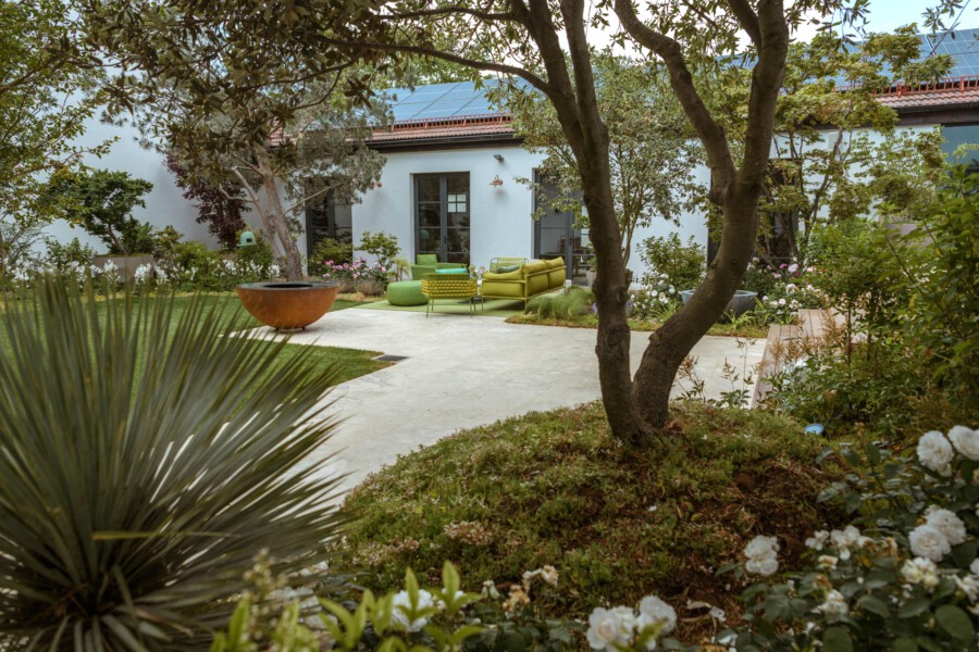 Gartenhof vor einem hellblauen Haus mit grüner Lounge-Möbel, Bäumen, blühenden Beeten und einem runden orangefarbenen Feuerkorb.