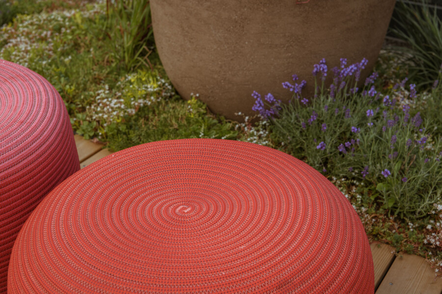 Rote, runde Sitzkissen mit geflochtenem Muster im Vordergrund; im Hintergrund Lavendelblüten, Grünpflanzen und ein großer brauner Pflanzkübel.