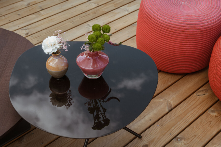 Glänzender runder Tisch mit zwei Vasen: links braun-beige Vase mit weißen Blüten, rechts rote Vase mit grünen Kugelblumen; Wolkenreflexe, rote Poufs und Holzdielen.