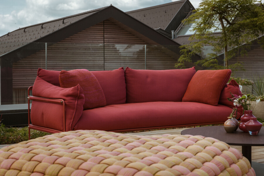Rotes Outdoor-Sofa mit Kissen auf Terrasse; vorne bunt geflochtener Sitzpouf, rechts Tisch mit Vasen, Hintergrund Glasbalustrade und Holzhaus.