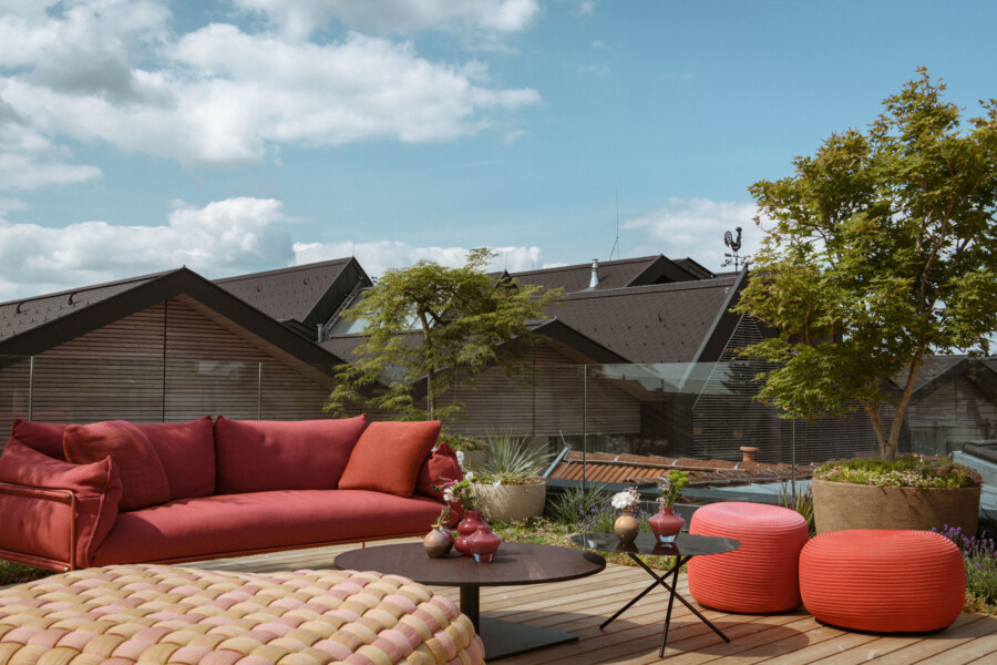 Dachterrasse mit rotem Loungesofa, roten Poufs, runden Beistelltisch und Pflanzkübeln; Glasbrüstung, Bäume und Häuserdächer vor blauem Himmel.
