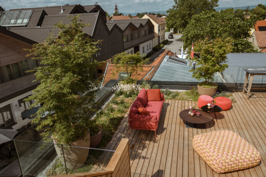 Holz-Dachterrasse mit rotem Sofa, roten Poufs und runden Tischen; Pflanzkübel, Glasgeländer, Blick auf Häuser und Bäume der Stadt.