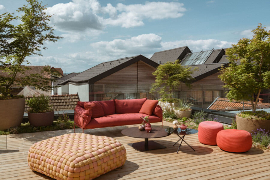 Rote Lounge auf einer Holzterrasse mit rundgeflochtener Sitzbank, zwei roten Poufs, Glastisch und Topfpflanzen; Blick auf Dächer und blauen Himmel.