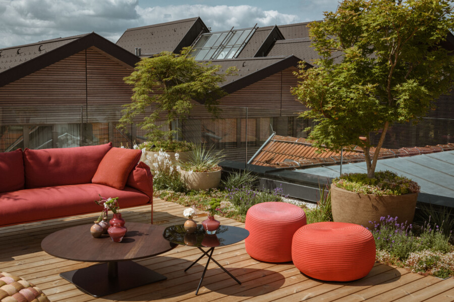 Rote Outdoor-Couch auf Holzterrasse mit zwei runden Poufs, Glastisch, Pflanzkübeln und Grünpflanzen; im Hintergrund moderne Dachhäuser bei Sonnenschein.