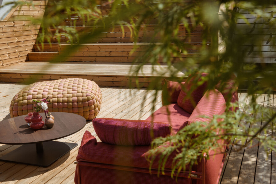 Holzdeck-Lounge im Freien mit rotem gepolstertem Sofa, rundem geflochtenem Hocker und Tisch mit Vasen und Blumen; grüne Blätter im Vordergrund.