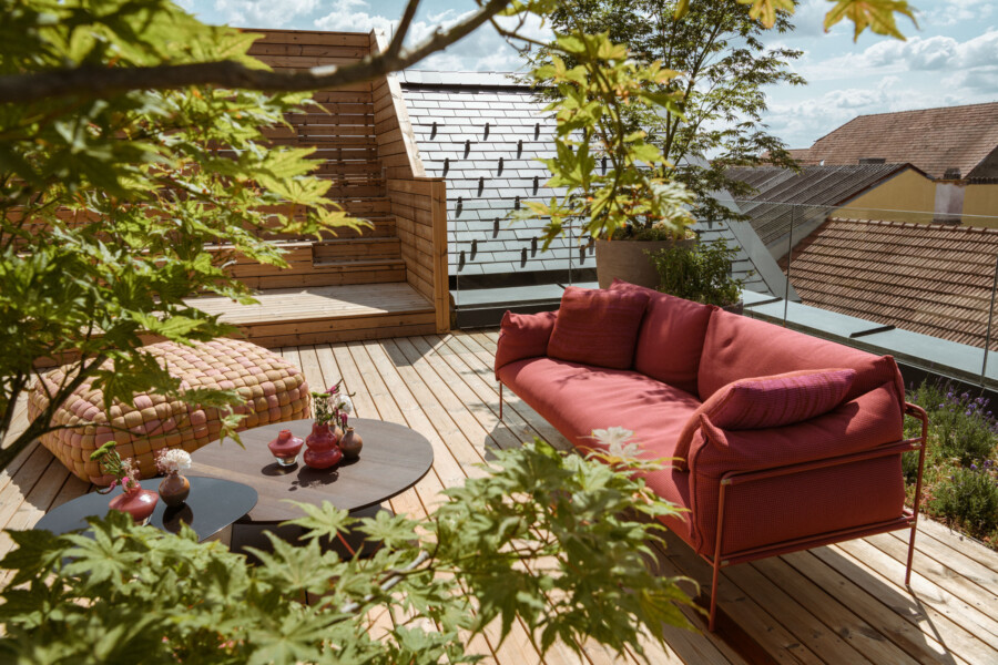 Rote Couch auf einer Holzterrasse mit Pouf, kleinem Tisch und Vasen, von grünem Laub umgeben; Glasbrüstung, Dächer und blauer Himmel im Hintergrund.