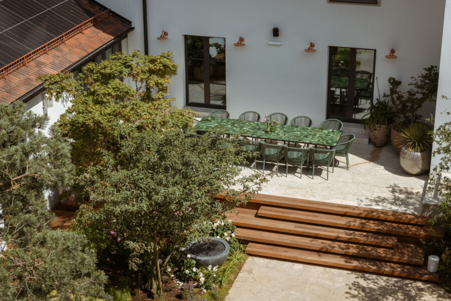 Blick von oben auf eine Terrasse: lange grüne Tischplatte mit Stühlen, umgeben von Topfpflanzen, helle Fassade, Holzstufen und Türen/Fenster.
