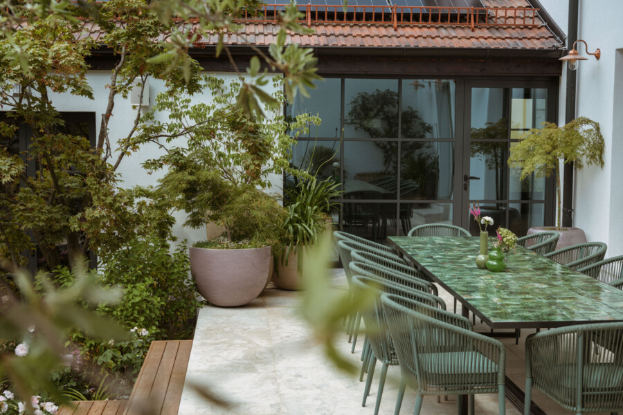 Terrasse mit langem Tisch aus grüner Marmorplatte und grünen Korbsesseln, von großen Topfpflanzen umgeben; Glas-Schiebetüren, rotes Ziegeldach oben.