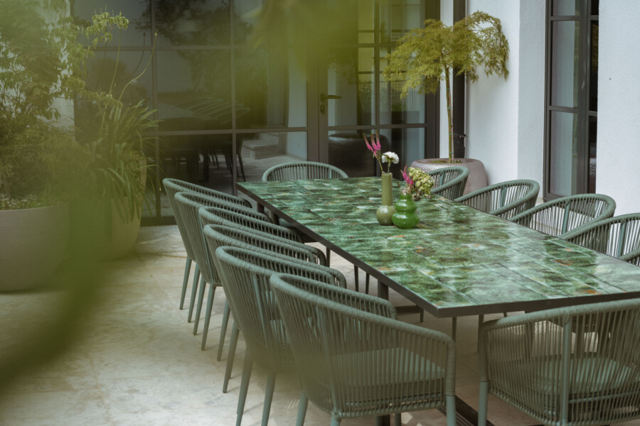 Außenbereich mit langem grünen Marmortisch und mehreren grünen Korbwickerstühlen; Vasen mit Blumen in der Tischmitte, Glas-Türen dahinter.