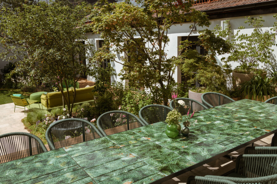 Garten mit langer grün marmorierter Esstischplatte und Korbgeflechtstühlen; dahinter grüne Sitzecke, Bäume und Blumentöpfe vor weißem Haus.