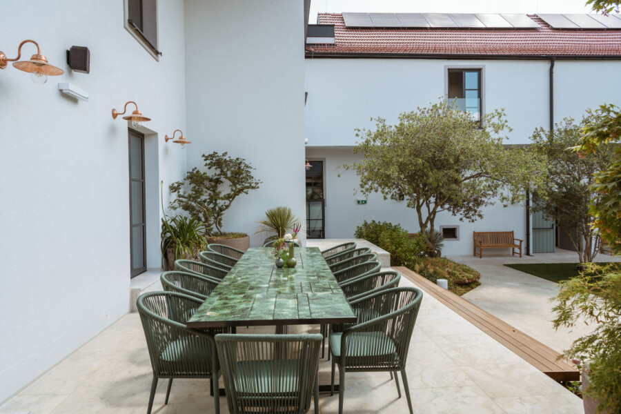 Außenhof eines modernen Gebäudes: langer grüner Marmortisch mit Geflechtstühlen, von Pflanzen umgeben, Wandlampen und eine Holzbank im Hintergrund.