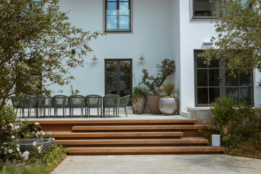 Außenansicht eines hellblauen Hauses mit Holzdeck und Stufen; Reihe grüner Stühle, Glas-Türen, sowie Topf- und Gartenpflanzen davor.