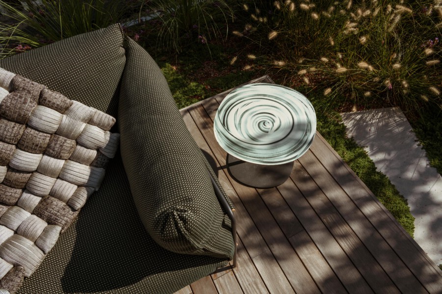 Outdoor wooden deck with a dark woven sofa and a chunky beige-cream woven pillow; a round glazed ceramic plate sits on a dark pedestal table, surrounded by ornamental grasses.