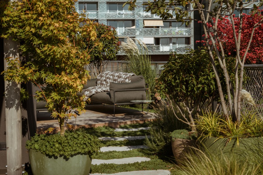 Sunny balcony garden with a brown outdoor sofa, quilted throw, glass side table, and lush potted plants; a modern apartment building in background.