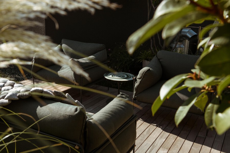 Outdoor seating on a wooden deck with dark green cushioned chairs around a round metal table with a spiral top, framed by lush plants.