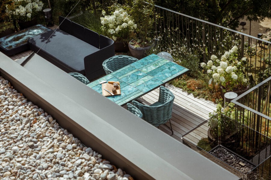 Außen-Terrasse von oben: türkisfarbener Fliesen-Tisch, zwei geflochtene Sessel, schwarzes Lounge-Sofa, Pflanzkübel mit weißen Hortensien am Metallgeländer, Kies links.