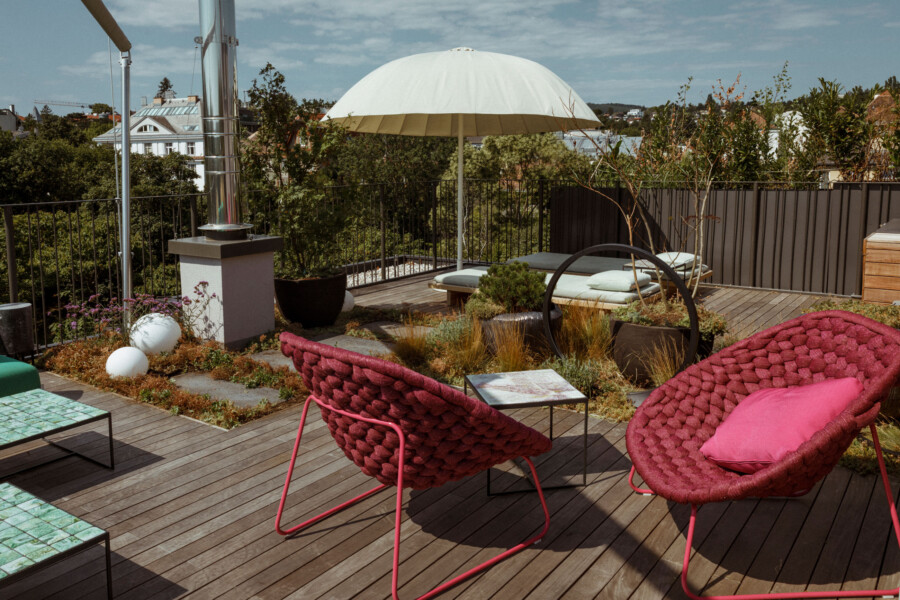 Dachterasse mit Holzdeck, weißem Sonnenschirm, zwei pink geflochtene Sessel mit Kissen, Beistelltisch, Pflanzen, Ring-Skulptur, Stadtblick.