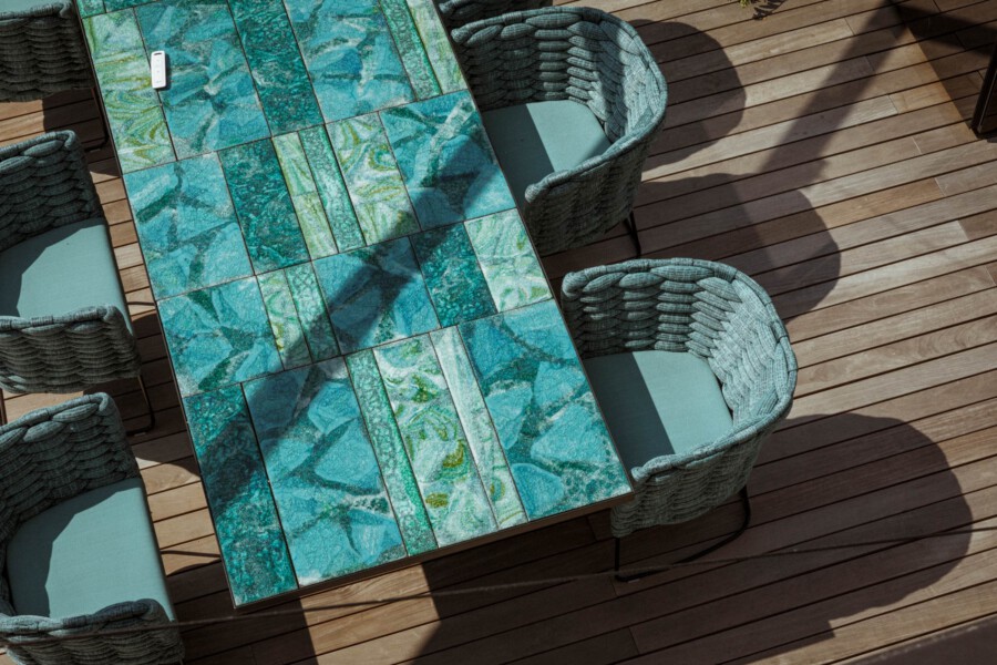 Vogelperspektive: Mosaiktisch mit blaugrünen Fliesen, umgeben von Korbstühlen mit blauen Kissen auf Holzdielen.