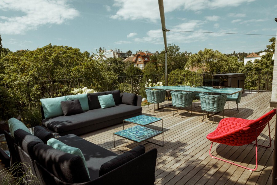 Eine Dachterrasse mit schwarzen Lounge-Sofas und türkisfarbenen Kissen, blauem Esstisch und gewebten Stühlen; roter Sessel, Holzdeck und grüner Stadtblick.