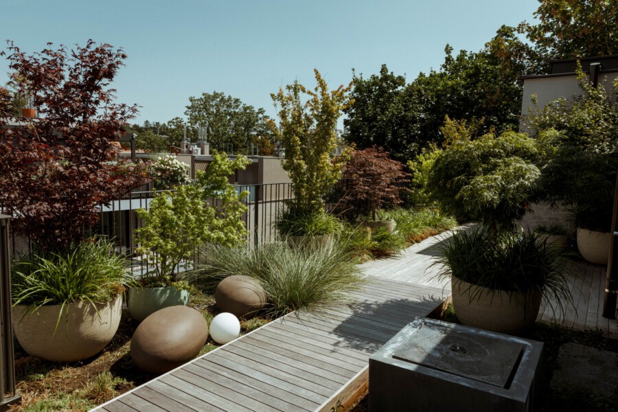 Dachgartenweg mit Holzplanken, großen Pflanzkübeln, Gräsern und buntem Laub; braune und weiße Kugeln, Metallgeländer, klarer blauer Himmel.
