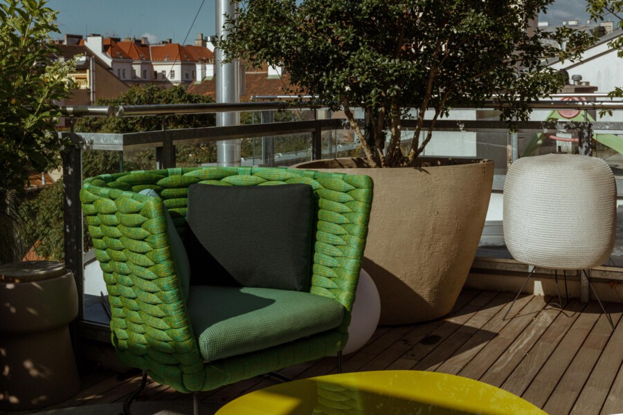 Green woven lounge chair with black cushion on a wooden rooftop terrace; large beige planter with Ficus sp. tree, glass railing, and white woven lamp.