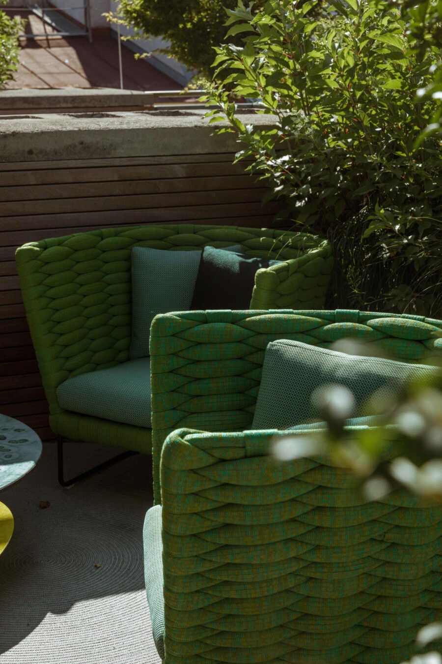 Patio seating with green woven rattan chairs; lush shrub with glossy leaves nearby (Latin name not identified).