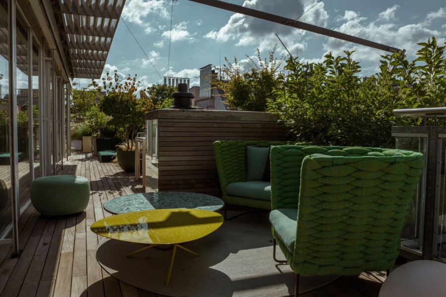 Rooftop terrace with green wicker seating around round tables, wooden deck, and potted shrubs along the railing, city skyline in the distance.