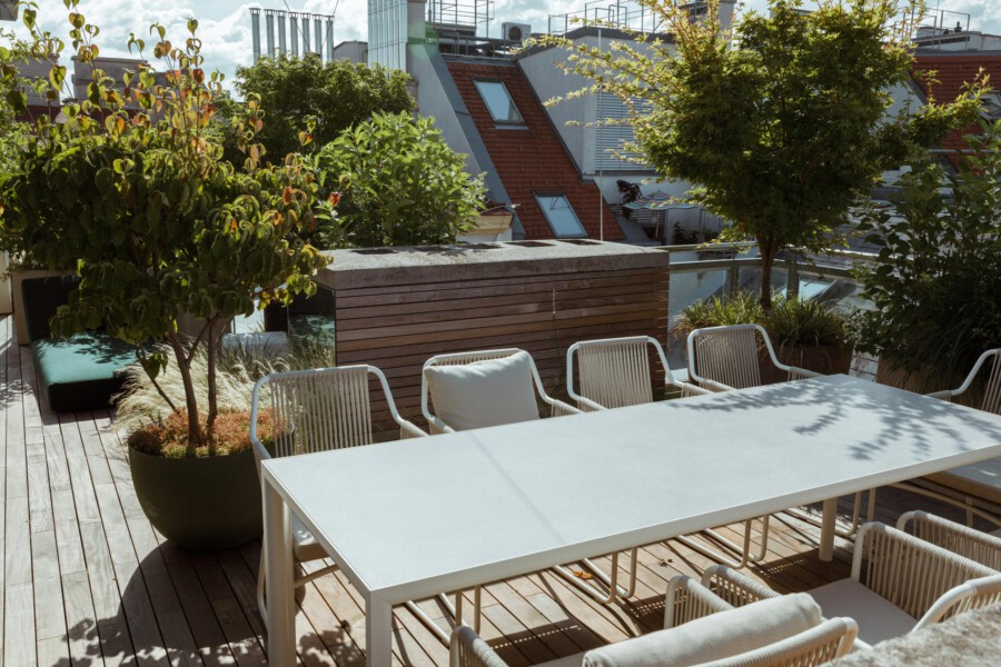 Rooftop terrace with a white rectangular dining table and white metal chairs, potted trees and grasses on a wooden deck.