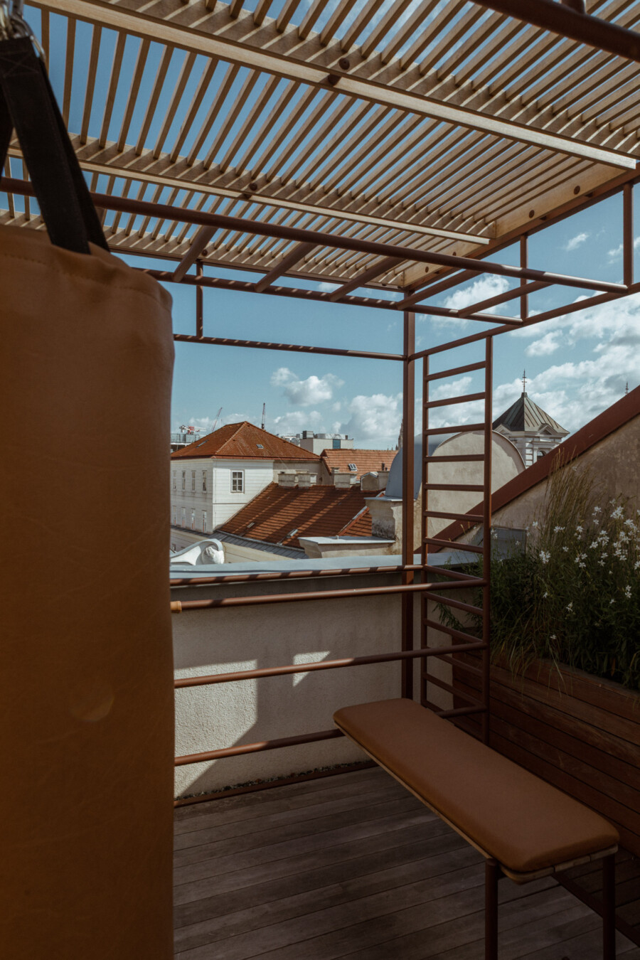 Dachterrasse mit braunem Boxsack links, brauner Sitzbank, Pflanzkasten mit Gräsern und weißen Blüten; Blick auf rote Dachziegeldächer der Stadt.
