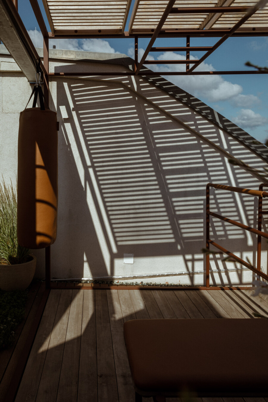 Hängender Boxsack vor Pergola-Veranda; Schatten der Lamellen auf Wand; Holzdeck; Topfpflanze Gräser (Pennisetum sp.).