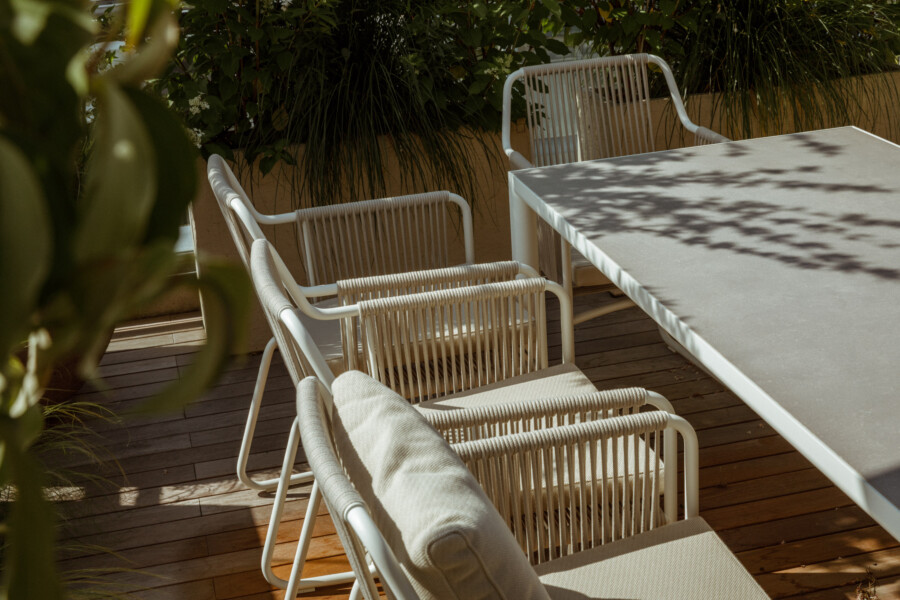 White woven patio chairs around a rectangular gray table on a wooden deck; tall ornamental grasses (Poaceae) and lush foliage in the background.