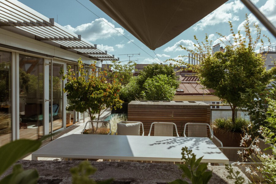 Rooftop terrace with a white table, metal chairs, potted trees, sliding glass doors, and a large shade sail.