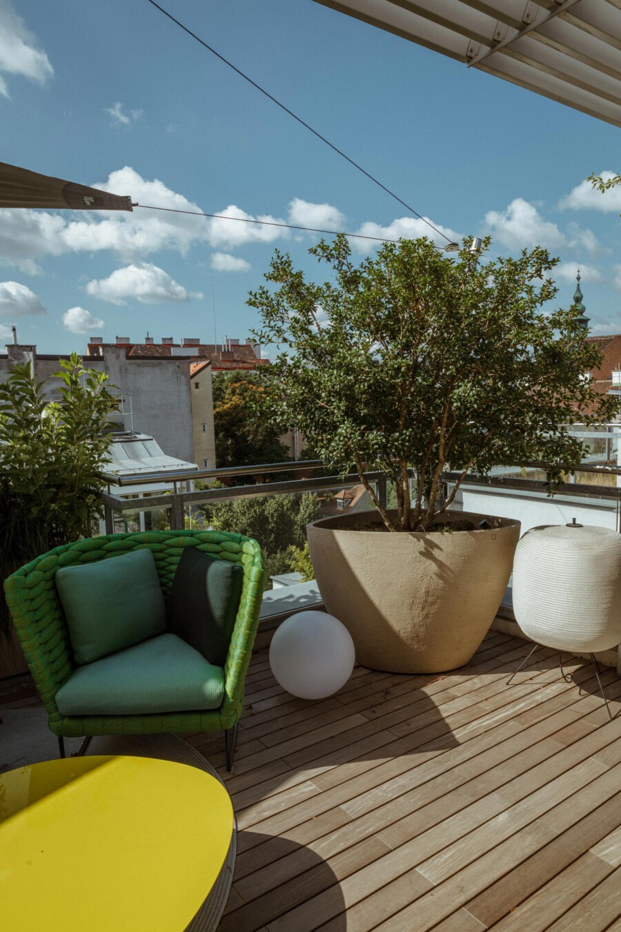 Ficus benjamina in a beige planter on a city balcony, with a green wicker chair, yellow table, and white spherical lamp.
